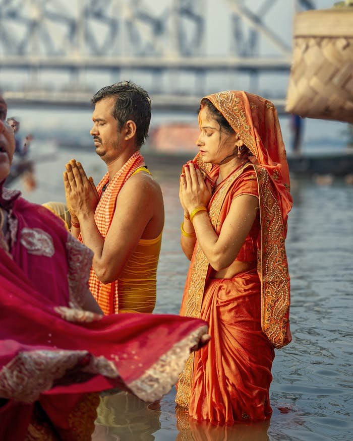 A couple in traditional attire performing Hindu rituals in a river, with the Howrah Bridge in the background.